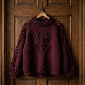 May include: A deep burgundy long-sleeve jumper featuring a prominent raised spider motif on the front. The garment has a ribbed mock neck, open lattice-style crochet detailing along the shoulders and sleeves, and a frayed hemline at the bottom.