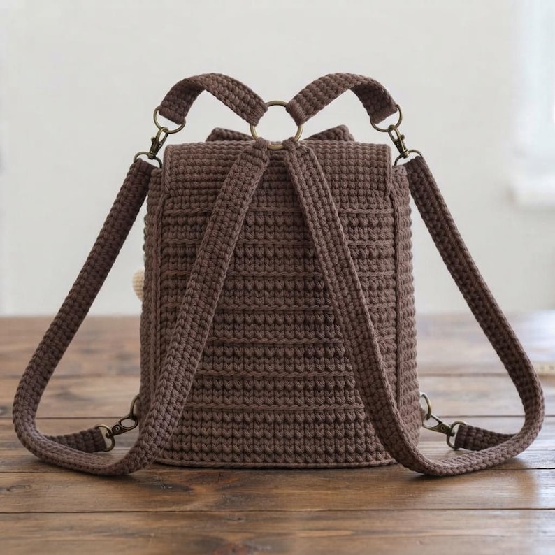 Peut inclure: Un sac &agrave; dos en crochet marron fonc&eacute; pr&eacute;sentant un motif de points c&ocirc;tel&eacute;s textur&eacute;s. Le sac comprend deux bretelles r&eacute;glables fix&eacute;es par des fermoirs pivotants en m&eacute;tal et un anneau m&eacute;tallique central sur le dessus, offrant un design structur&eacute;.