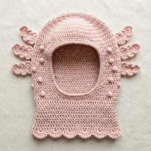 May include: A light pink crochet balaclava featuring a large central face opening, scalloped bottom edges, and three decorative branch-like appendages on each side. The surface is accented with small, raised bobble stitches throughout.