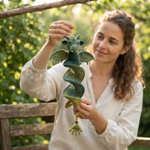 May include: A green crocheted dragon hanging ornament with a spiral-shaped body, small wings, and a head with two horns. The dragon features a gradient of green yarn shades and terminates in a small hand-shaped tail with a string of colourful beads.