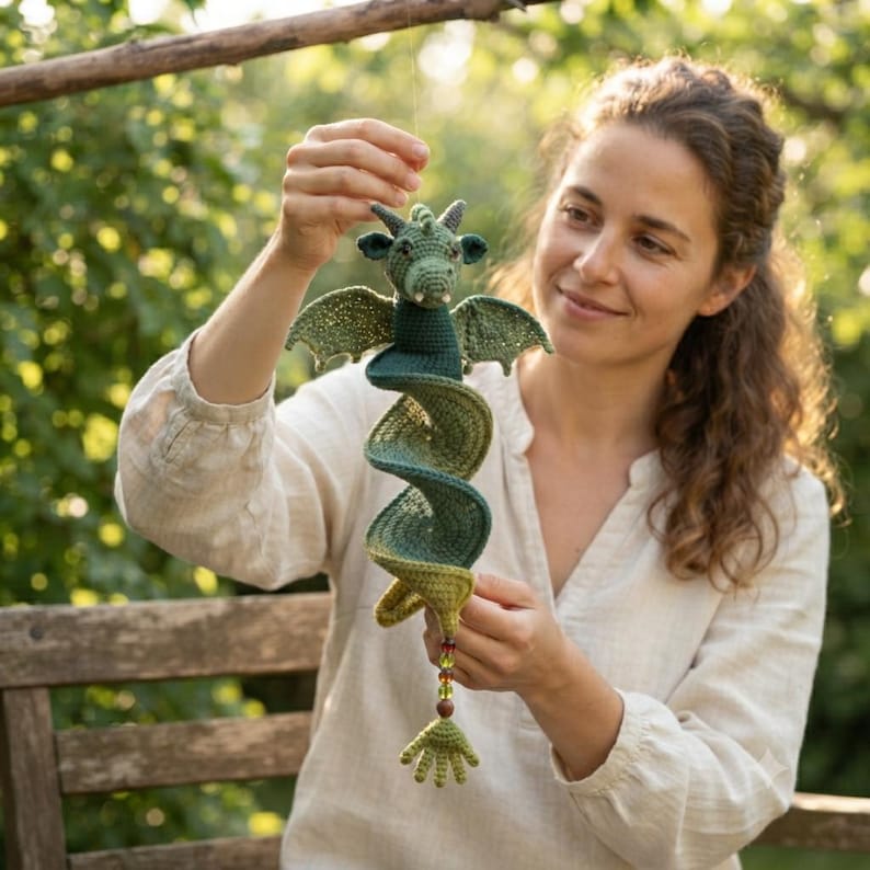 May include: A green crocheted dragon hanging ornament with a spiral-shaped body, small wings, and a head with two horns. The dragon features a gradient of green yarn shades and terminates in a small hand-shaped tail with a string of colourful beads.