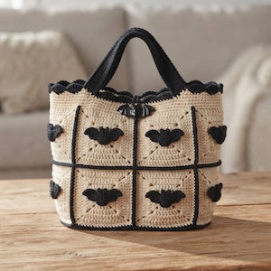 May include: A beige crochet tote bag constructed from square panels with black trim and handles. The bag features black crocheted bat appliques centered on each square panel and a small black bat charm attached to the top center edge.