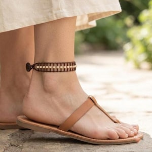 May include: A brown anklet featuring a woven cord design with small cream-colored beads arranged in a row, secured with a dark brown button closure. The anklet is worn above a tan leather thong sandal with a simple strap across the foot.