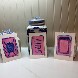 May include: Three watercolor paintings with pink and blue designs. The paintings are on white paper and feature a floral design, Chinese characters, and a geometric pattern. The paintings are displayed in front of two blue and white ceramic jars.