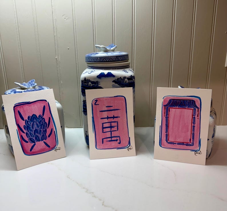 May include: Three watercolor paintings with pink and blue designs. The paintings are on white paper and feature a floral design, Chinese characters, and a geometric pattern. The paintings are displayed in front of two blue and white ceramic jars.