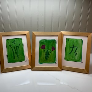 May include: Three framed art pieces with gold-colored frames. Each piece features a green background with a unique design: a stylized "W", two purple flowers, and the number "9" with a symbol. The art is on white paper.