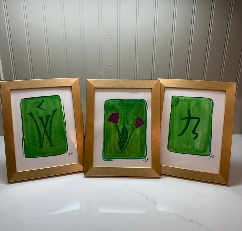 May include: Three framed art pieces with gold-colored frames. Each piece features a green background with a unique design: a stylized "W", two purple flowers, and the number "9" with a symbol. The art is on white paper.