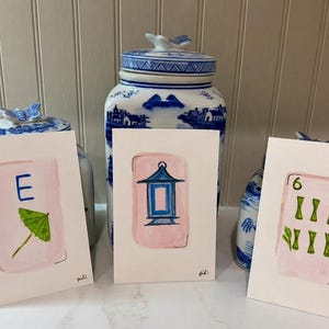 May include: Three watercolor paintings of Mahjong tiles. Each tile is on a white background with a pink border. One tile has the letter "E" and a green umbrella. Another has a blue lantern. The third has the number "6" and green bamboo.