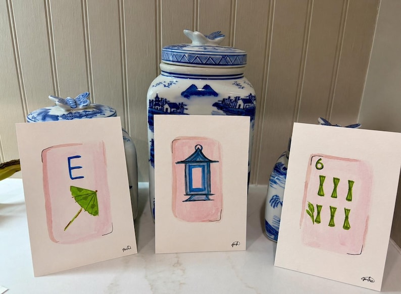 May include: Three watercolor paintings of Mahjong tiles. Each tile is on a white background with a pink border. One tile has the letter "E" and a green umbrella. Another has a blue lantern. The third has the number "6" and green bamboo.