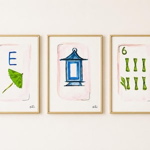 May include: Three framed watercolor prints with gold frames. Each print features a design on a white background with a pink wash. The designs include a blue letter "E" and green umbrella, a blue lantern, and the number "6" with green bamboo.