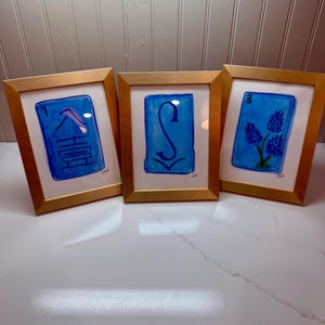 May include: Three framed watercolor paintings with gold frames. Each painting features a blue background with a white border. The paintings display a number, a symbol, and a floral design. The frames are displayed on a white surface.