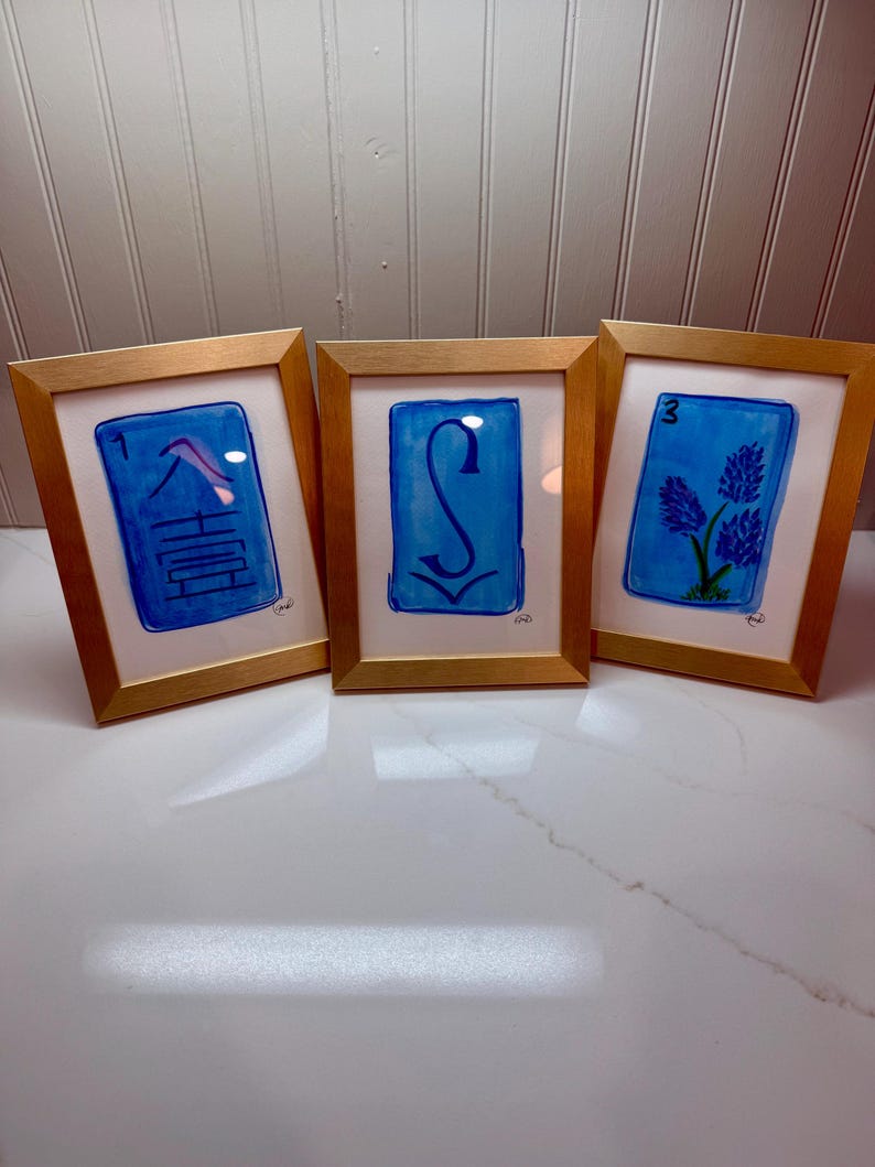 May include: Three framed watercolor paintings with gold frames. Each painting features a blue background with a white border. The paintings display a number, a symbol, and a floral design. The frames are displayed on a white surface.