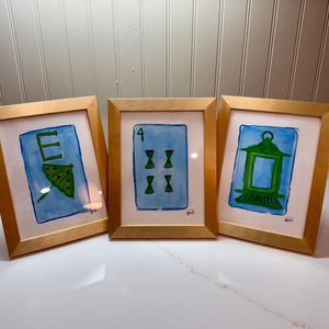 May include: Three framed watercolor paintings with gold frames. The paintings feature blue backgrounds with green and white illustrations of a letter, a playing card, and a lantern. The frames are displayed on a white surface.