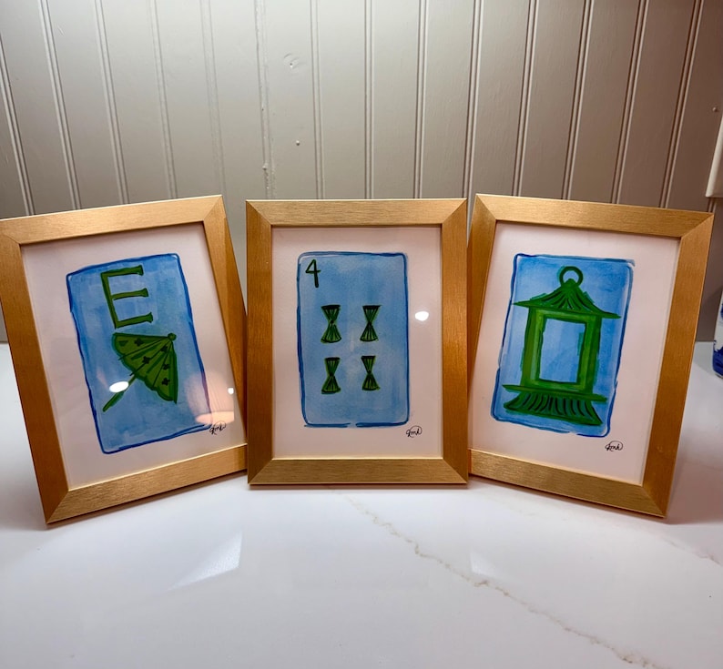 May include: Three framed watercolor paintings with gold frames. The paintings feature blue backgrounds with green and white illustrations of a letter, a playing card, and a lantern. The frames are displayed on a white surface.