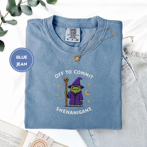 May include: A blue jean coloured t-shirt featuring an embroidered graphic of a green frog wearing a purple wizard hat and robe, holding a wooden staff. The text reads OFF TO COMMIT SHENANIGANS, surrounded by small yellow stars and a crescent moon.