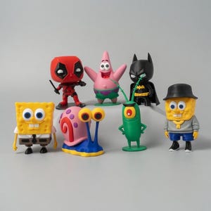 May include: A collection of colorful figurines featuring characters from popular cartoons and comic books. The figures include Deadpool, Patrick Star, Batman, SpongeBob SquarePants, Gary the Snail, and Plankton. Each figure is uniquely designed and painted.