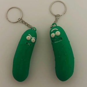May include: Two green pickle-shaped keychains with cartoon faces, silver key rings, and chain attachments. The keychains have white eyes with red pupils and are set against a white background.