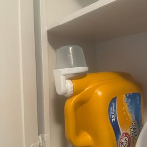 May include: A yellow laundry detergent container with a white dispenser attached. The container has a handle and a red cap. The dispenser is clear and has a measuring cup shape. The product is stored in a white cabinet.