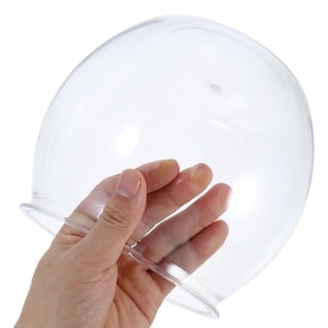May include: A clear, dome-shaped glass cloche with a flared base. The cloche is held by a hand, showcasing its transparency and smooth surface. Ideal for displaying small items or protecting delicate objects.
