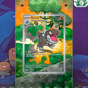 May include: A Tyrunt Pokémon card with a brown and red dinosaur illustration, set against a green and orange jungle background. The card is displayed in a clear acrylic holder. Text on the card includes "Tyrunt" and "Get Angry".
