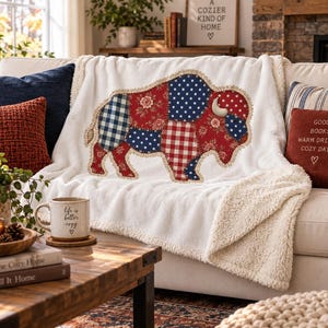 May include: A white plush throw blanket featuring a large central applique of a buffalo. The buffalo is constructed from various fabric patches in red, white, and blue, showcasing patterns including gingham, polka dots, and floral prints.