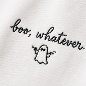 Op de afbeelding: Een wit stoffen oppervlak met zwarte borduursels. De tekst "boo, whatever." is in cursief schrift geborduurd. Daaronder staat een kleine, zwart geborduurde omtrek van een geest met de armen omhoog in een schouderophalend gebaar.
