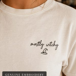 May include: An off-white crew neck t-shirt featuring black embroidery on the left chest. The text reads mostly witchy in a cursive script, accompanied by a small embroidered witch hat icon below the text. The shirt has a ribbed neckline.