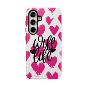 Peut inclure: Une coque de smartphone blanche ornée d'un motif répétitif de cœurs rose vif à l'aspect brossé. Le texte "Wife Life" est imprimé en écriture cursive noire au centre, accompagné de deux petits cœurs noirs de chaque côté du mot life.