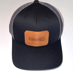 May include: A black baseball cap with a grey mesh back and a rectangular tan leather patch on the front. The patch features the letters GMC embossed in the centre. The cap has a solid black front panel and a curved brim with visible stitching.