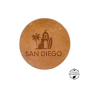 May include: A circular tan leather coaster featuring a dark brown laser-engraved design of a palm tree, a mission-style building with a bell, and a surfboard. The text SAN DIEGO is engraved below the graphic. A small logo reads REINVENTED BOARDS.