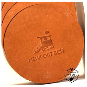 Op de afbeelding: Een stapel ronde, bruinleren onderzetters. Elke onderzetter heeft een reliëfontwerp van een strandwachttoren met de tekst "NEWPORT BCH". De onderzetters hebben een warme, aardse tint en een eenvoudig ontwerp.
