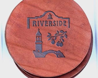 May include: A round brown leather coaster featuring a debossed design with the word RIVERSIDE, a bell icon, a tower, a bridge, and a leafy branch with fruit. The coaster has a smooth texture and a dark brown finish on the edges.