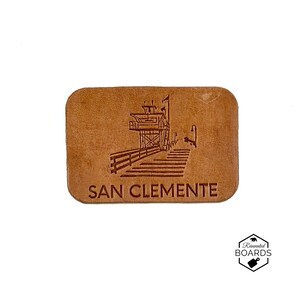 May include: A rectangular tan leather patch featuring a dark brown laser-engraved illustration of the San Clemente pier and lifeguard tower. The text SAN CLEMENTE is printed in capital letters below the pier graphic on the leather surface.