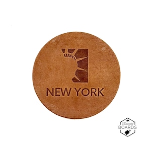 Op de afbeelding: Een ronde onderzetter van bruin leer met een donkerbruine reliëfafbeelding van de kroon en stralen van het Vrijheidsbeeld in een vierkant kader, met de tekst NEW YORK in hoofdletters onder de afbeelding op het oppervlak gedrukt.
