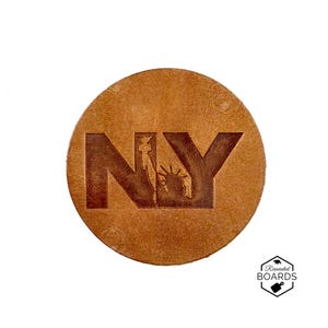 Op de afbeelding: Een ronde, lichtbruine leren patch met de letters NY in reliëf in een donkerbruine kleur. De letter Y bevat een gedetailleerd silhouet van het Vrijheidsbeeld. Het oppervlak heeft een natuurlijke nerfstructuur en een gladde, matte afwerking.