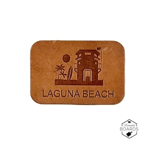Puede incluir: Un parche rectangular de cuero color canela con un diseño en relieve de una torre de salvavidas, palmeras, una tabla de surf y un sol. El texto LAGUNA BEACH está grabado en mayúsculas debajo del gráfico. Se incluye un logo REINVENTED BOARDS.