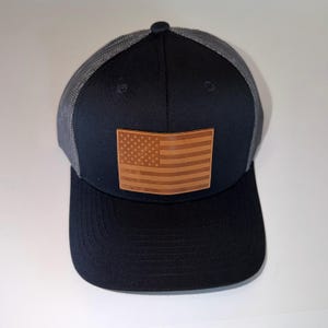 Könnte beinhalten: Eine schwarze Baseballkappe mit grauer Netzrückseite und flachem Schirm. Auf der Vorderseite befindet sich ein rechteckiger, brauner Lederaufnäher, der mit dem Design der amerikanischen Flagge inklusive Sternen und Streifen geprägt ist.