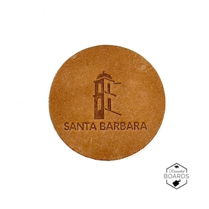 May include: A circular tan leather coaster featuring a dark brown embossed graphic of a mission-style bell tower and the text SANTA BARBARA printed in a sans-serif font below the tower. The coaster has a smooth, matte finish and clean edges.
