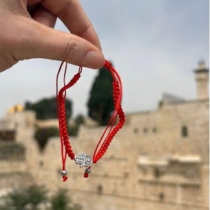 May include: Red string bracelet with a silver Hamsa hand charm. The bracelet is made of braided red string and features a small silver charm in the shape of a hand with intricate details. The bracelet is held up against a blurred outdoor background.
