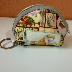 May include: A semi-circular fabric pouch featuring a vintage-style collage print with teddy bears, roses, and French text. The pouch has a grey fabric trim, a top zipper closure, and a silver-toned metal keyring attached to the side.