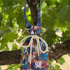 Puede incluir: Una bolsa con cordón hecha de tela de patchwork con una variedad de patrones florales y de olas en azul, rojo, amarillo y verde. La bolsa tiene un asa de tela única y un cierre de cuerda grueso de color crema con puntas plateadas.