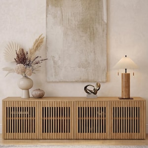 Könnte beinhalten: Ein helles Holzschrank mit vertikalen Lamellentüren, gekrönt von einer Lampe, einer abstrakten Skulptur und einer Vase mit getrockneten Blumen. Ein großes abstraktes Gemälde hängt an der Wand. Ein Teppich befindet sich im Vordergrund.