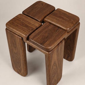 Könnte beinhalten: Ein Hocker aus dunkelbraunem Holz mit einem einzigartigen Design. Er hat vier quadratische Sitze, die miteinander verbunden sind. Die geschwungenen Beine sorgen für Stabilität. Dieses Holzmöbel eignet sich für jeden Raum. Maße: ca. 40 cm x 40 cm.