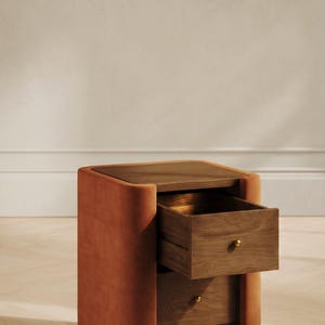 Könnte beinhalten: Ein moderner Nachttisch mit zwei Schubladen. Der Rahmen ist mit einem rostfarbenen Stoff bezogen, und die Oberseite und die Schubladen sind aus Holz. Eine Schublade ist geöffnet und gibt den Blick ins Innere frei.