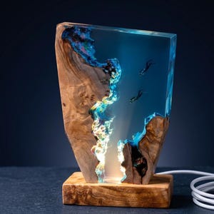 May include: A unique wooden lamp with a clear resin center, featuring a blue ocean scene with divers. The lamp has a rectangular wooden base and a natural wood grain exterior. The resin is illuminated from within, creating a glowing effect.