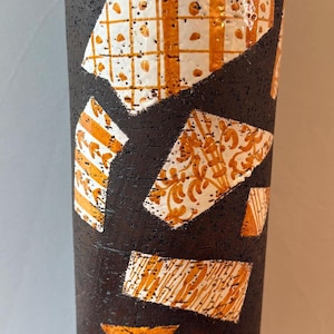 May include: A tall cylindrical vase with a dark brown, textured matte finish. The surface is decorated with several white and orange geometric patches featuring various patterns, including grids, floral motifs, and linear textures.