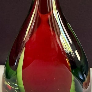 May include: A teardrop-shaped glass sculpture featuring a clear base that transitions into a vibrant green layer, topped with a deep red core. The smooth, polished surface reflects light, highlighting the layered colour composition and the solid, rounded form of this decorative glass object.