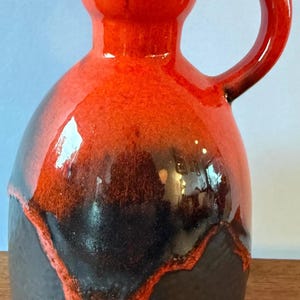 May include: A ceramic pitcher with a glossy finish, featuring a vibrant orange upper section that transitions into a dark, textured black base. The vessel has a rounded body, a narrow neck, and a single curved handle extending from the side.
