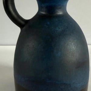 May include: A ceramic pitcher with a rounded body and a single handle. The vessel features a dark brown base that transitions into a mottled blue glaze on the upper portion, ending in a smooth, narrow neck with a circular opening at the top.