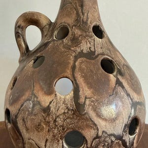 May include: A round ceramic vase with a narrow neck and a single side handle. The surface features a marbled brown and tan glaze with multiple circular cutouts distributed across the body, creating a hollow, open-work design throughout the piece.